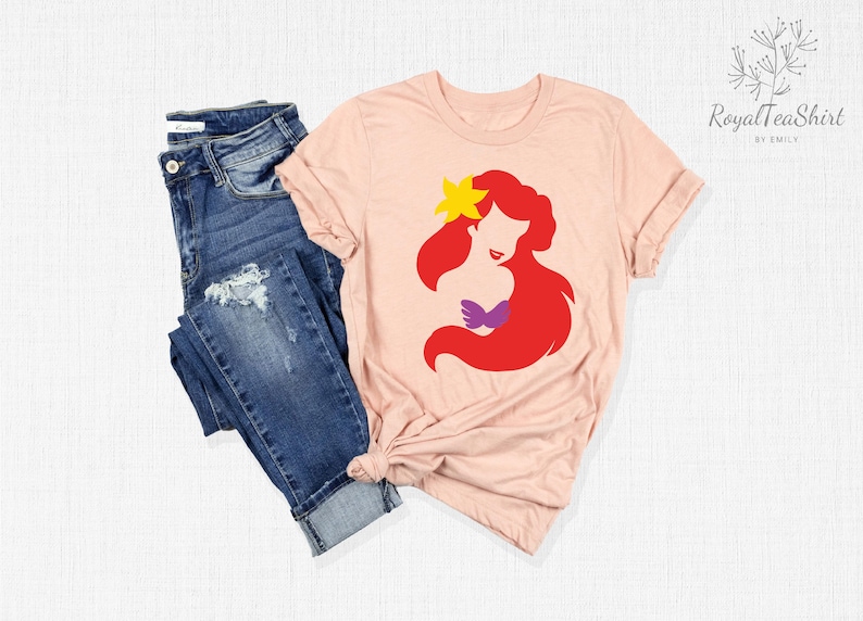 May include: A pink t-shirt with a silhouette of Ariel, the Little Mermaid, in red, purple, and yellow. The shirt is displayed on a white surface with a pair of blue jeans.