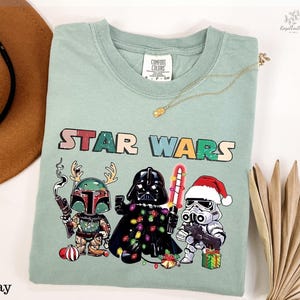 A green t-shirt with a Star Wars-themed graphic design featuring three characters: a character with a gun, a character with a Christmas stocking, and a character with a gift box. The t-shirt is displayed on a white background, along with a hat, a necklace, and some decorative elements.