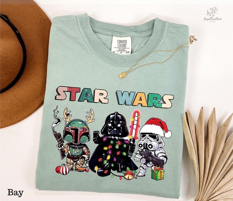 A green t-shirt with a Star Wars-themed graphic design featuring three characters: a character with a gun, a character with a Christmas stocking, and a character with a gift box. The t-shirt is displayed on a white background, along with a hat, a necklace, and some decorative elements.