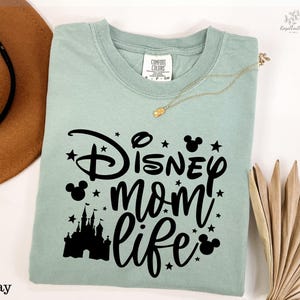 A light green t-shirt with a Disney-themed design, including a castle silhouette and stars, along with the text "Disney mom life" printed on it. The t-shirt is laid out on a white surface, with a brown hat and a fan visible in the background.