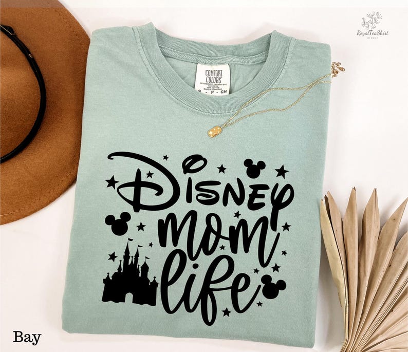 A light green t-shirt with a Disney-themed design, including a castle silhouette and stars, along with the text "Disney mom life" printed on it. The t-shirt is laid out on a white surface, with a brown hat and a fan visible in the background.