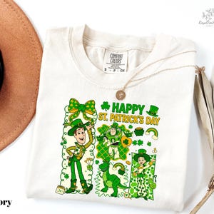 A white t-shirt with a cartoon design of St. Patrick's Day characters, including a man, a woman, and a child, along with various St. Patrick's Day symbols and text.