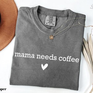 A gray t-shirt with the text "mama needs coffee" printed on it, along with a heart symbol, is displayed on a white background.