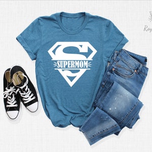 K&ouml;nnte beinhalten: Ein blaues T-Shirt mit einem wei&szlig;en Superman-Logo und dem Wort "SUPERMOM" darauf gedruckt. Das Shirt liegt flach auf einer wei&szlig;en Oberfl&auml;che mit einem Paar schwarzer Turnschuhe und einer blauen Jeans.