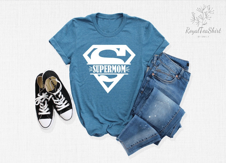 K&ouml;nnte beinhalten: Ein blaues T-Shirt mit einem wei&szlig;en Superman-Logo und dem Wort "SUPERMOM" darauf gedruckt. Das Shirt liegt flach auf einer wei&szlig;en Oberfl&auml;che mit einem Paar schwarzer Turnschuhe und einer blauen Jeans.