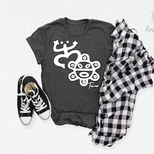 May include: A dark gray t-shirt with a white graphic of a stylized frog and a sun with a face. The text "Taino" is below the sun. The shirt is laid flat on a white surface with a black and white plaid shirt and black sneakers.