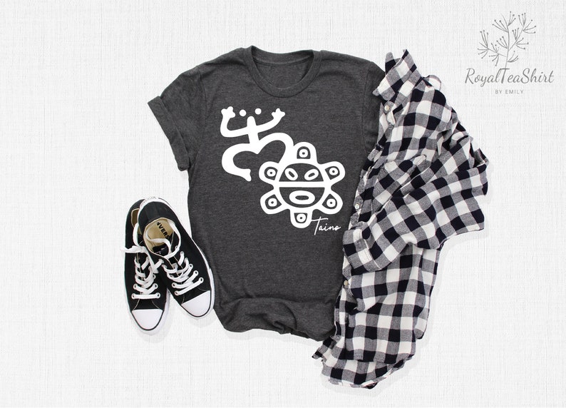 May include: A dark gray t-shirt with a white graphic of a stylized frog and a sun with a face. The text "Taino" is below the sun. The shirt is laid flat on a white surface with a black and white plaid shirt and black sneakers.