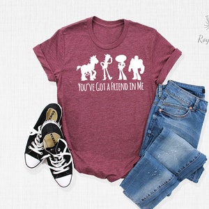 May include: A maroon t-shirt featuring a white silhouette of Woody, Buzz Lightyear, and Jessie from Toy Story. The text "You've got a friend in me" is printed below the silhouettes. The shirt is laid out on a white surface with a pair of black Converse shoes and a pair of blue jeans.