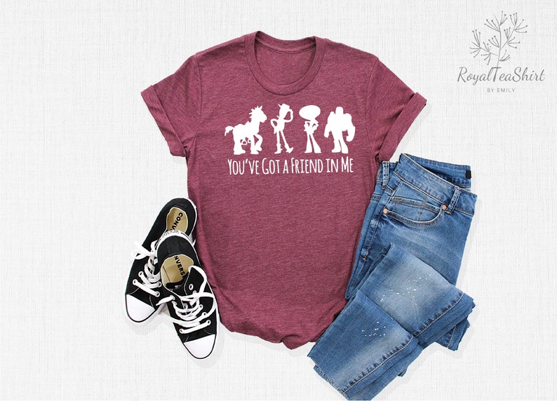 May include: A maroon t-shirt featuring a white silhouette of Woody, Buzz Lightyear, and Jessie from Toy Story. The text "You've got a friend in me" is printed below the silhouettes. The shirt is laid out on a white surface with a pair of black Converse shoes and a pair of blue jeans.