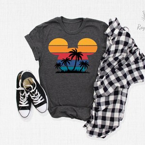 May include: A dark gray t-shirt with a retro sunset design in the shape of Mickey Mouse ears. The design features palm trees and a blue ocean.