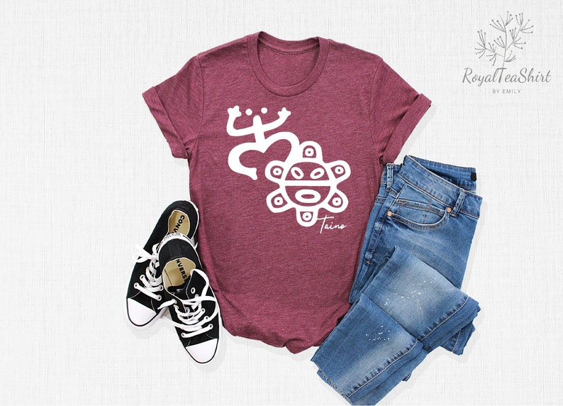 May include: A maroon t-shirt with a white design of a stylized frog and sun with the word "Taino" underneath. The shirt is laid flat on a white surface with a pair of black sneakers and blue jeans.