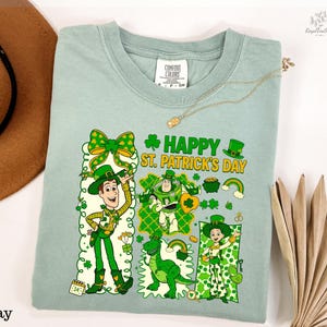 A light green t-shirt with a cartoon design of characters from the Toy Story franchise, including Woody and Buzz Lightyear, along with St. Patrick's Day-themed elements. The t-shirt is displayed on a white background, accompanied by a hat, a necklace, and some leaves.