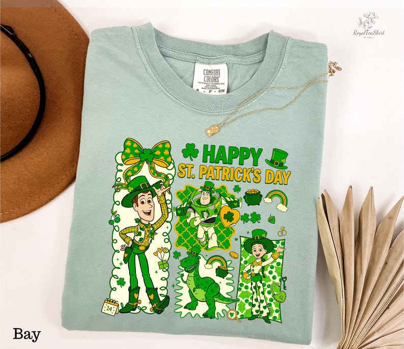 A light green t-shirt with a cartoon design of characters from the Toy Story franchise, including Woody and Buzz Lightyear, along with St. Patrick's Day-themed elements. The t-shirt is displayed on a white background, accompanied by a hat, a necklace, and some leaves.