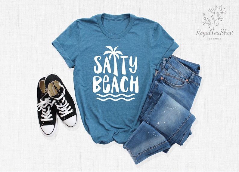 May include: A teal blue t-shirt with a white graphic that says "SAPPY BEACH" with a palm tree and waves. The shirt is laid out on a white surface with a pair of black sneakers and blue jeans.