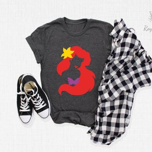May include: A dark grey t-shirt with a red silhouette of Ariel from The Little Mermaid. The silhouette includes a purple seashell bra and a yellow flower in her hair.
