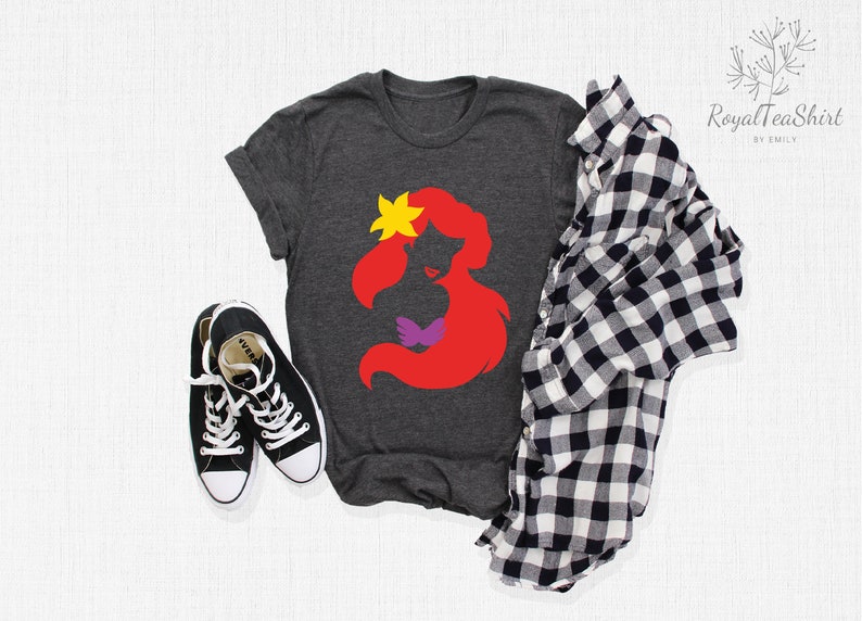 May include: A dark grey t-shirt with a red silhouette of Ariel from The Little Mermaid. The silhouette includes a purple seashell bra and a yellow flower in her hair.