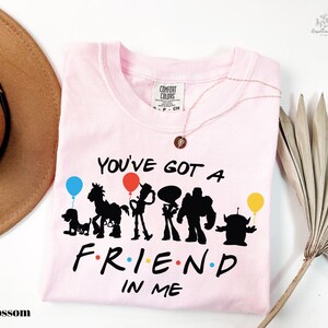 A pink t-shirt with a graphic design featuring silhouettes of characters from the TV show Toy Story, along with the text "You've Got a Friend in Me". The t-shirt is displayed on a white background, accompanied by a brown hat and a palm frond.