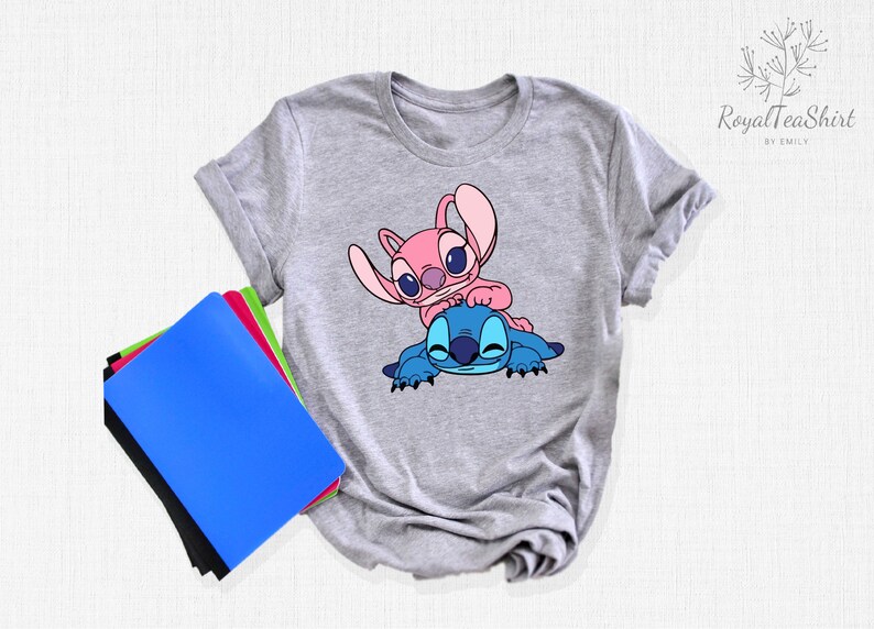 K&ouml;nnte beinhalten: Graues T-Shirt mit einem blauen und rosa Stitch- und Angel-Cartoon-Design. Das Design zeigt Angel auf Stitch.