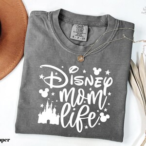 A gray t-shirt with a Disney-themed design, including a castle and stars, along with the text "Disney mom life" printed on it. The t-shirt is folded and placed on a white surface, with a brown hat and some leaves nearby.