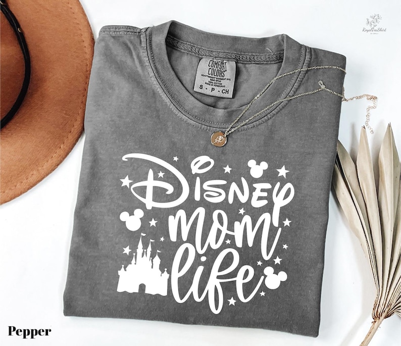 A gray t-shirt with a Disney-themed design, including a castle and stars, along with the text "Disney mom life" printed on it. The t-shirt is folded and placed on a white surface, with a brown hat and some leaves nearby.