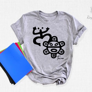 May include: A gray t-shirt with a black graphic of a stylized sun with a face and a frog-like figure. The text "Taino" is printed below the graphic.