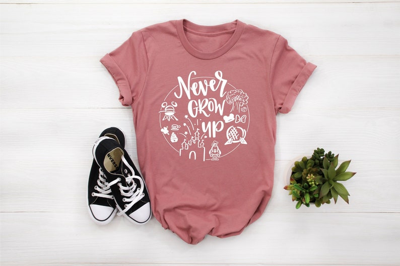 May include: A pink t-shirt with a white circle graphic that says "Never Grow Up" and includes various Disney park icons.