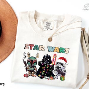 A white t-shirt with a colorful graphic design featuring characters from the Star Wars franchise, including Darth Vader, Stormtrooper, and an elf.