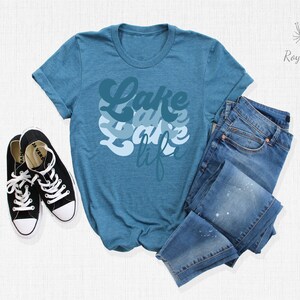 May include: A teal blue t-shirt with the text "Lake Life" in a layered, retro font design. The shirt is laid flat on a white surface with a pair of black sneakers and blue jeans.