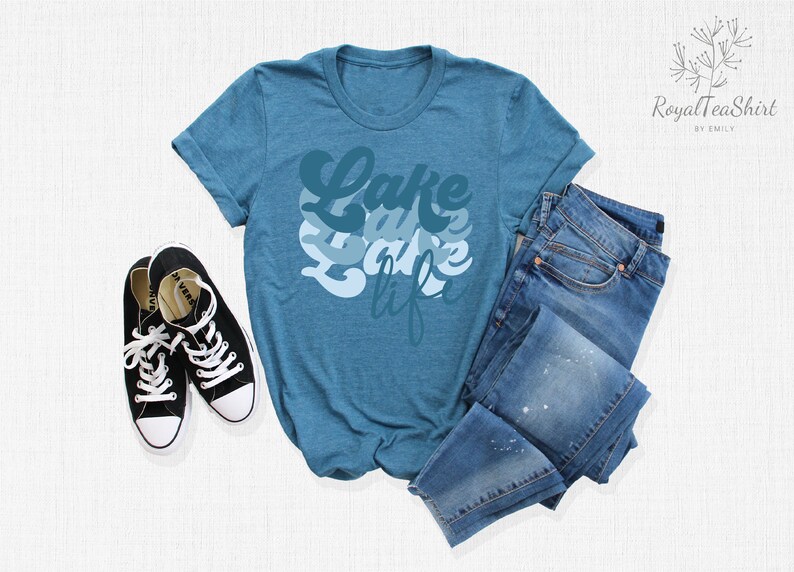 May include: A teal blue t-shirt with the text "Lake Life" in a layered, retro font design. The shirt is laid flat on a white surface with a pair of black sneakers and blue jeans.