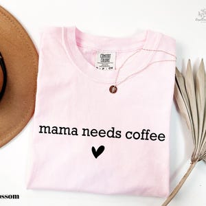 A pink t-shirt with the text "mama needs coffee" printed on it, along with a heart symbol, is displayed alongside a brown hat, a necklace, and a palm frond.