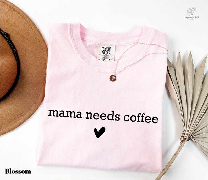 A pink t-shirt with the text "mama needs coffee" printed on it, along with a heart symbol, is displayed alongside a brown hat, a necklace, and a palm frond.
