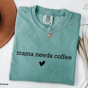 A light green t-shirt with the text "mama needs coffee" printed on it, along with a heart symbol. The t-shirt is folded and placed on a white surface, accompanied by a brown hat, a necklace, and some dried leaves.
