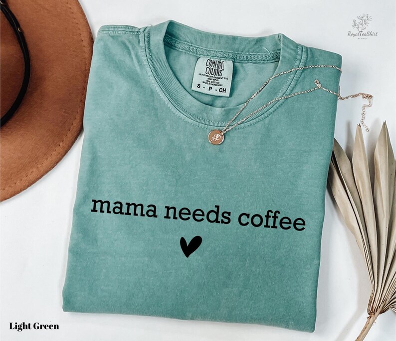 A light green t-shirt with the text "mama needs coffee" printed on it, along with a heart symbol. The t-shirt is folded and placed on a white surface, accompanied by a brown hat, a necklace, and some dried leaves.