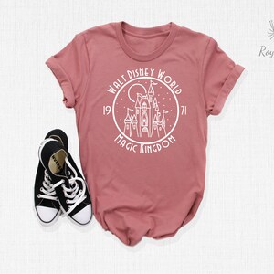 May include: A dusty rose t-shirt with a white graphic of Cinderella's castle in a circle with the text "Walt Disney World 1971 Magic Kingdom".