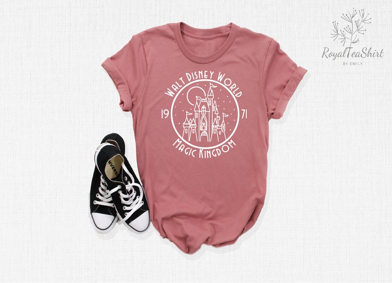 May include: A dusty rose t-shirt with a white graphic of Cinderella's castle in a circle with the text "Walt Disney World 1971 Magic Kingdom".