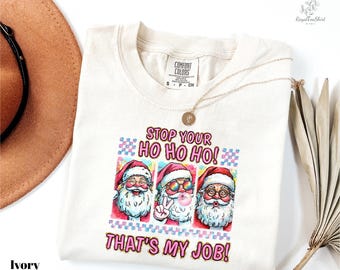 May include: Ivory-colored t-shirt with a graphic of three Santa Clauses, each with a different expression. The text "STOP YOUR HO HO HO! THAT'S MY JOB!" is printed in pink and blue. A gold necklace is draped over the shirt.
