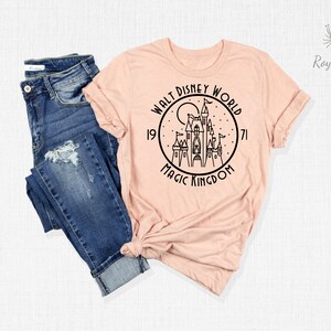 May include: A peach-colored t-shirt with a black graphic of Cinderella's castle in a circle with the text "Walt Disney World 1971 Magic Kingdom".