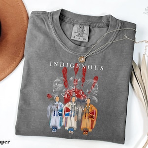 May include: A gray Comfort Colors t-shirt with the word "INDIGENOUS" printed on the front. The graphic features Native American figures and a red handprint design. A silver necklace with a pendant is draped across the shirt.