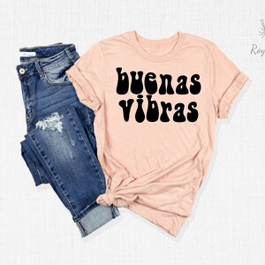 May include: A light pink t-shirt with the text "buenas vibras" in black retro-style lettering. The shirt is shown on a white background with a pair of blue jeans.