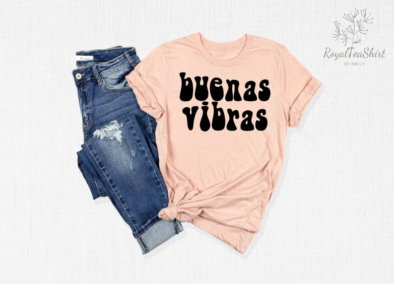 May include: A light pink t-shirt with the text "buenas vibras" in black retro-style lettering. The shirt is shown on a white background with a pair of blue jeans.