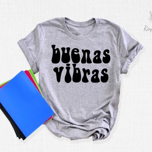 May include: A gray t-shirt with the text "buenas vibras" in black, retro-style lettering. The shirt is laid flat on a white surface with a stack of notebooks to the left.