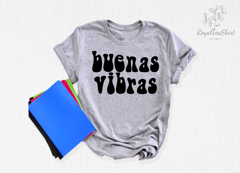 May include: A gray t-shirt with the text "buenas vibras" in black, retro-style lettering. The shirt is laid flat on a white surface with a stack of notebooks to the left.