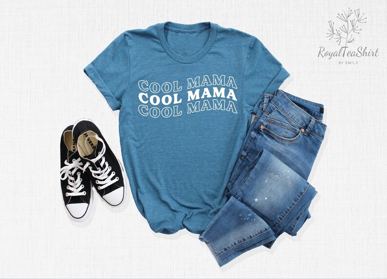 May include: A teal blue t-shirt with the text "COOL MAMA" repeated three times in white outlined text. The shirt is laid flat on a white surface with a pair of black sneakers and blue jeans.