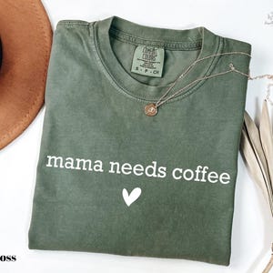 A green t-shirt with the text "mama needs coffee" printed on it, along with a heart symbol, is displayed on a white background.