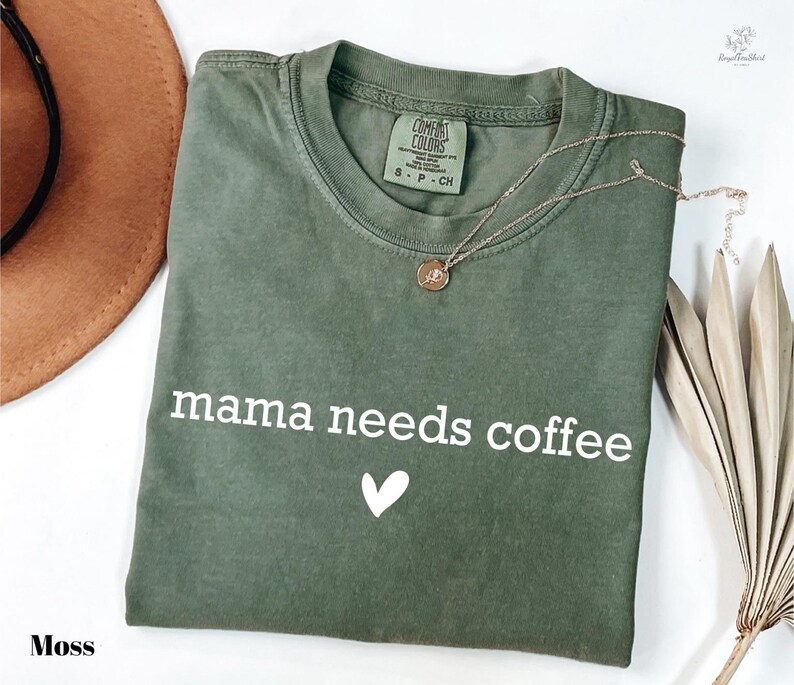 A green t-shirt with the text "mama needs coffee" printed on it, along with a heart symbol, is displayed on a white background.