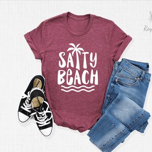 May include: A maroon t-shirt with a white graphic that says "SAPPY BEACH" with a palm tree and waves. The shirt is laid flat on a white surface with a pair of black sneakers and blue jeans.