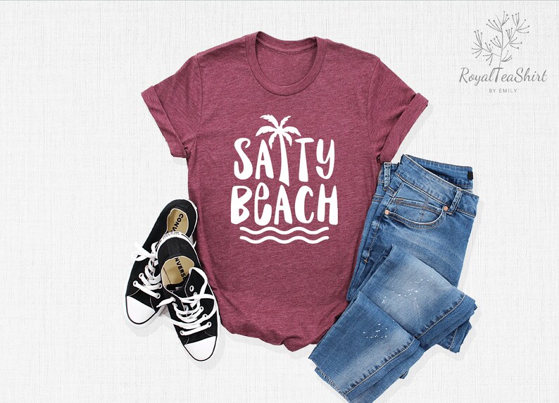 May include: A maroon t-shirt with a white graphic that says "SAPPY BEACH" with a palm tree and waves. The shirt is laid flat on a white surface with a pair of black sneakers and blue jeans.