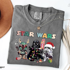 A gray t-shirt with a Star Wars-themed graphic design featuring three characters: a character in a green suit, a character in a red suit, and a character in a white suit. The characters are holding various Star Wars-themed items, such as a Star Wars box and a Star Wars peace sign. The t-shirt is displayed on a white surface, along with a brown hat and some dried leaves.