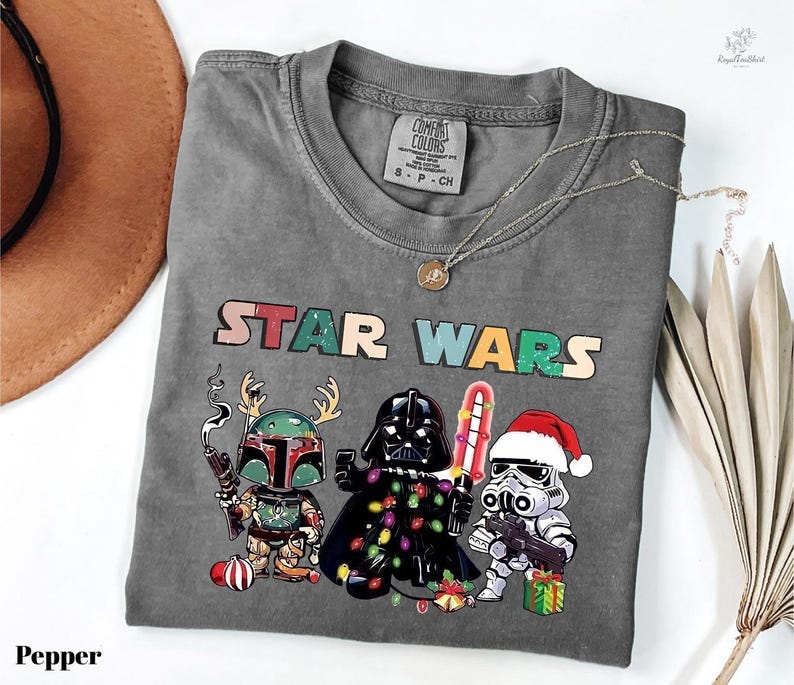 A gray t-shirt with a Star Wars-themed graphic design featuring three characters: a character in a green suit, a character in a red suit, and a character in a white suit. The characters are holding various Star Wars-themed items, such as a Star Wars box and a Star Wars peace sign. The t-shirt is displayed on a white surface, along with a brown hat and some dried leaves.