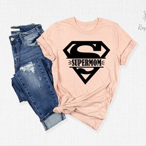 K&ouml;nnte beinhalten: Ein pfirsichfarbenes T-Shirt mit einem schwarzen Superman-Logo und dem Text "SUPERMOM" im Logo.